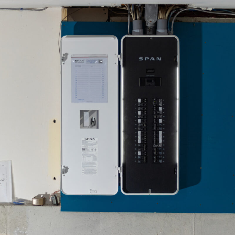 A SPAN smart electrical panel installed next to a traditional circuit breaker in a garage, with visible wiring at the top.