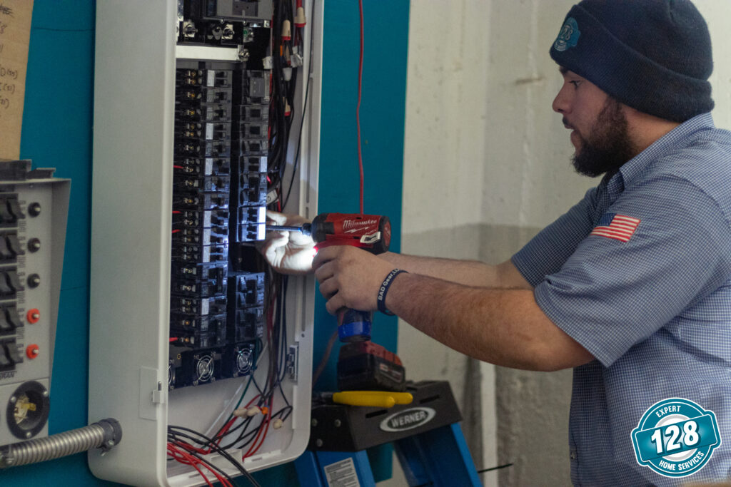 An electrician using a drill to work on an open electrical panel, wearing a beanie and a shirt with a usa flag patch.