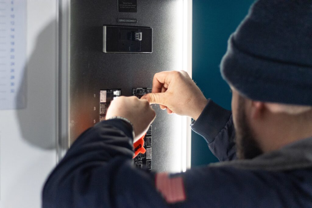 A person wearing dark clothing and a beanie adjusts a breaker switch on an electrical panel.