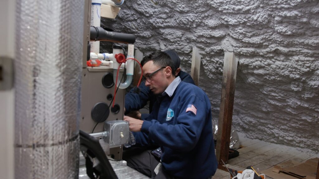 A technician wearing glasses and a blue jacket works on equipment in a spray foam insulated room.
