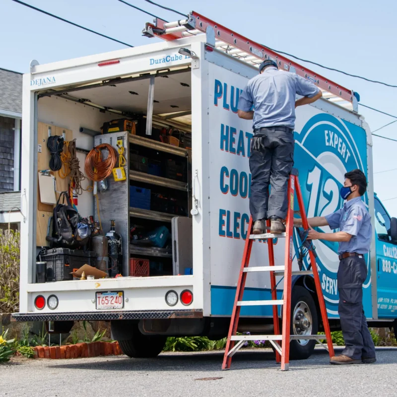 Two workers in uniform organize equipment in the back of a plumbing and heating service truck. One worker stands inside the truck, and the other adjusts a ladder. The truck has various tools and supplies.