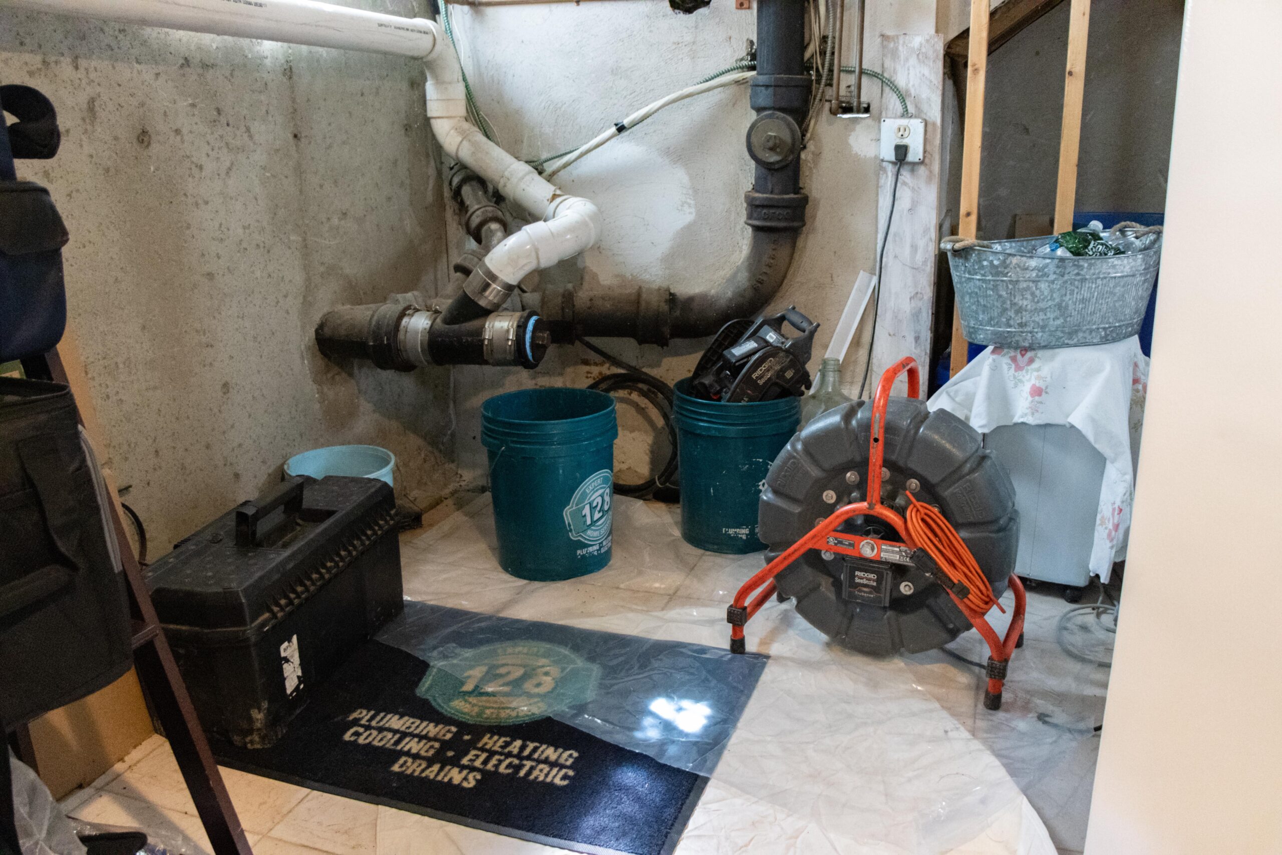 A basement utility area with plumbing pipes, a tool box, a mat, several buckets, and an electric drain cleaner. A shelf with a tablecloth and a metal bin are visible on the right.