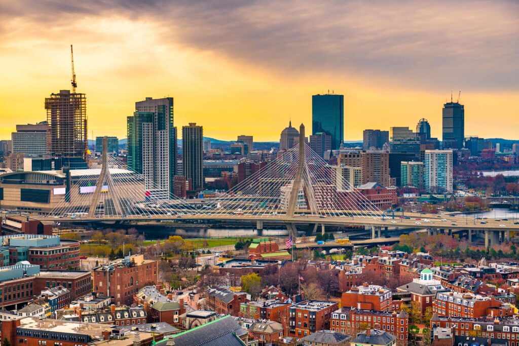 Aerial view of a city skyline with skyscrapers, a cable-stayed bridge, and a mix of modern and historic buildings under a yellow and orange sky.