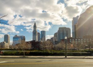 Urban scene featuring a cityscape with a clock tower, various skyscrapers, and bare trees against a blue sky with clouds. A road and sidewalk are in the foreground.