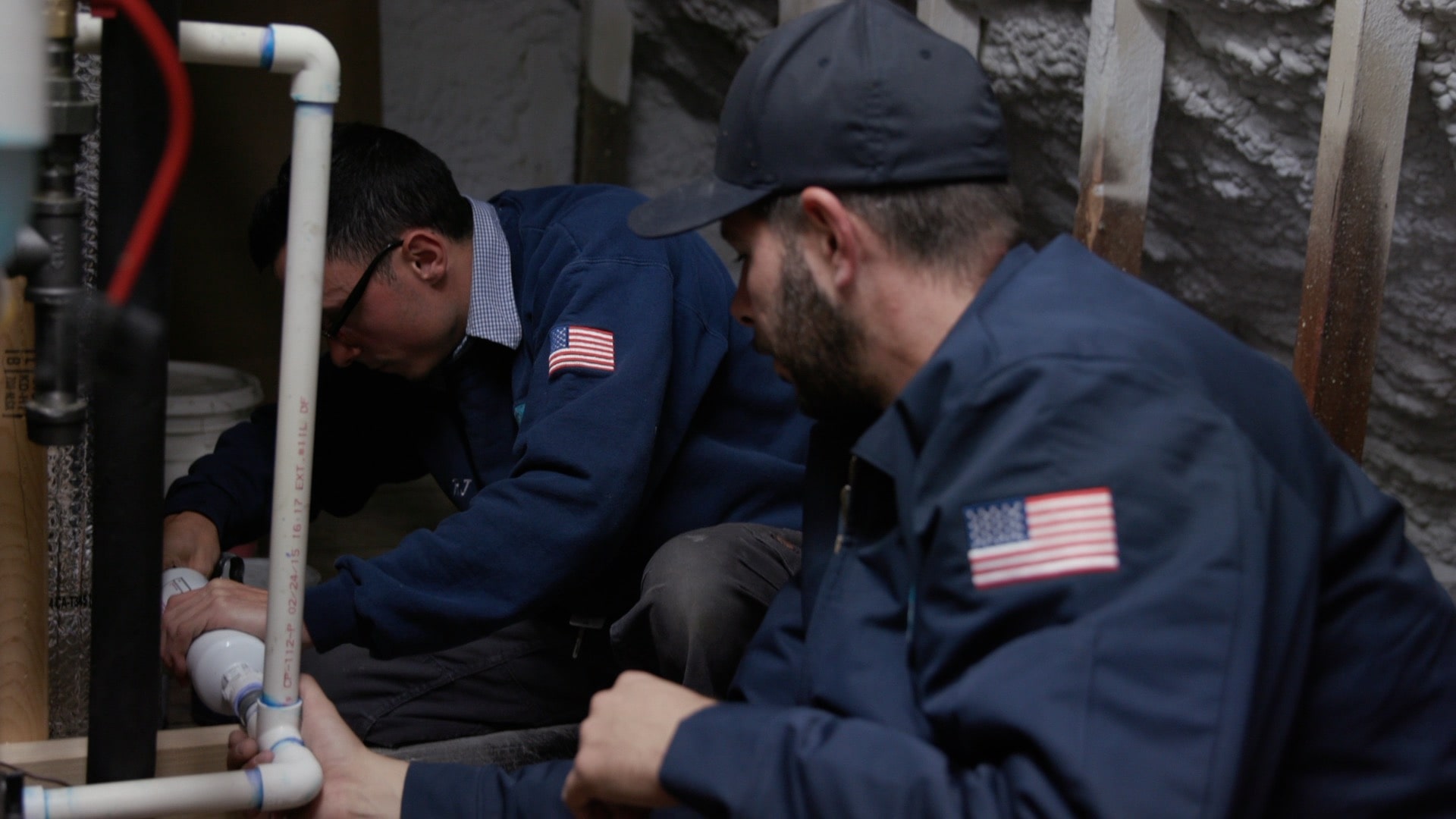 Two workers in blue uniforms with American flag patches perform plumbing tasks in a confined space with various pipes.