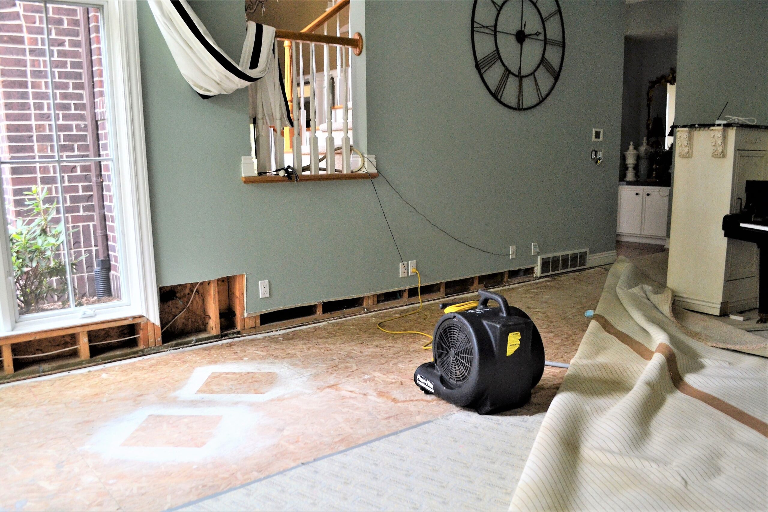 Room with stripped floor and walls, exposed studs, and a large fan for drying. Carpet is rolled up and furniture is covered; a staircase with a hammock in the background.