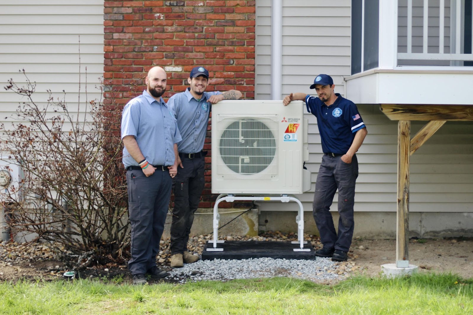 Three men in work uniforms stand next to an outdoor air conditioning unit mounted on a stand beside a building.