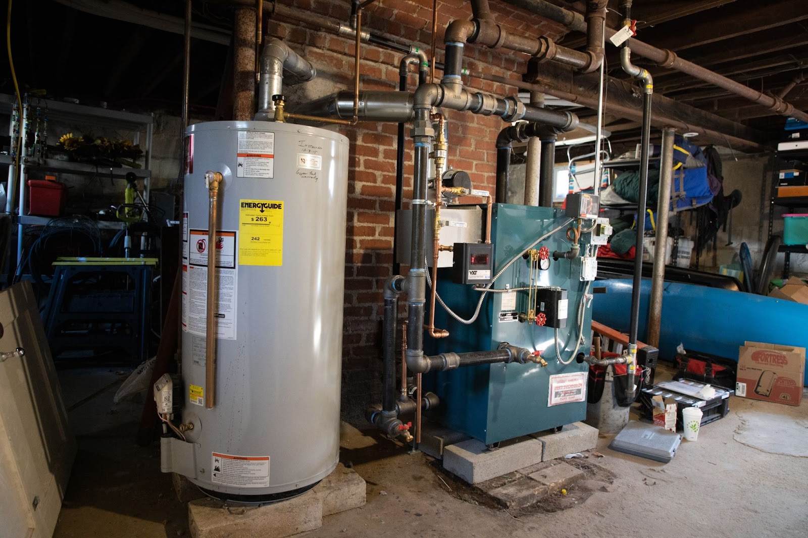 Basement with water heater and furnace connected by pipes. Tools and storage items around, brick wall in background.