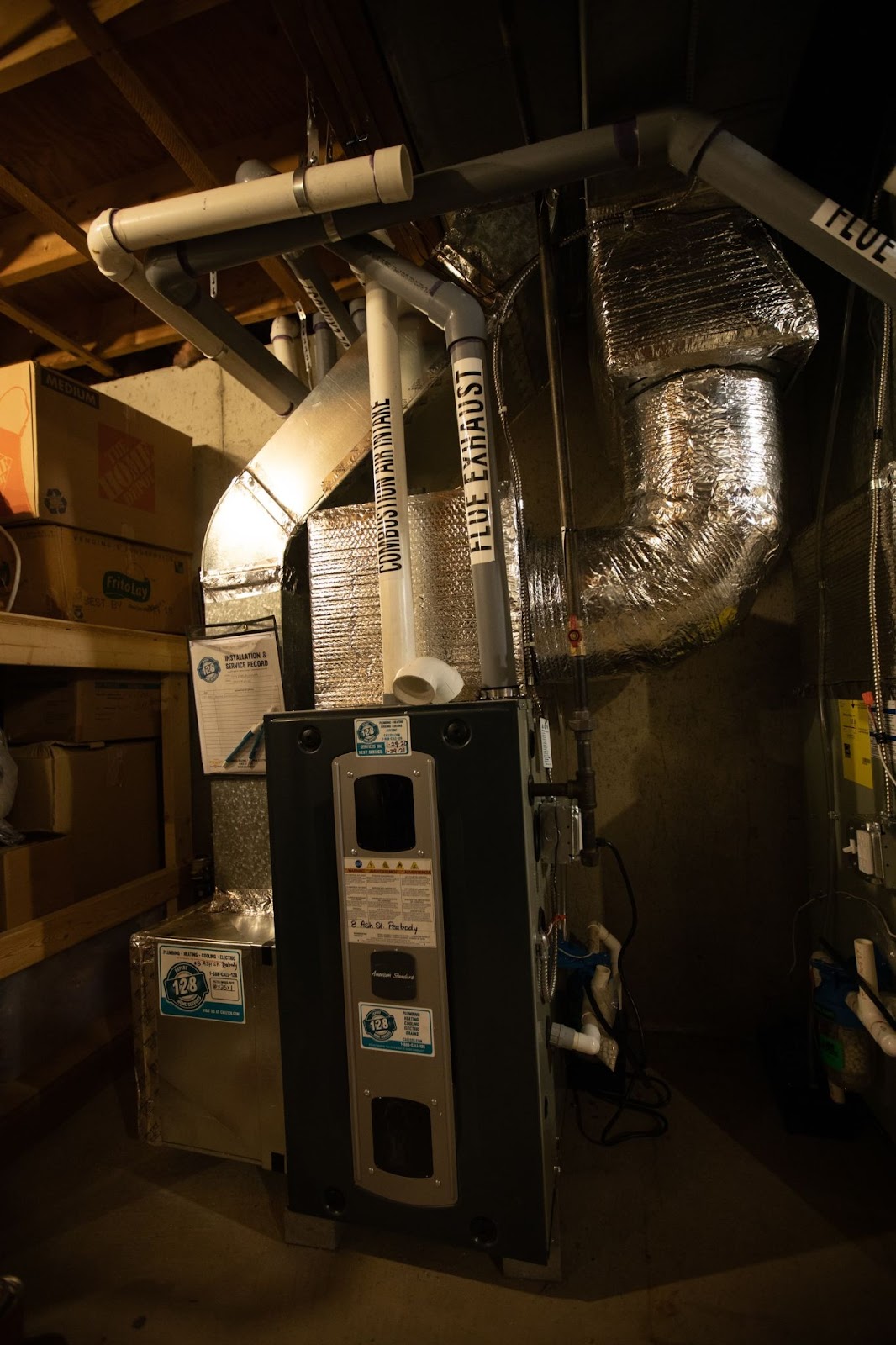 A basement with a central heating system, showing a furnace connected to an array of ducts and pipes. Shelves with boxes and various storage items are visible on the left.