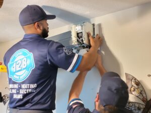 Two technicians install an air conditioning unit on a wall. One is adjusting the device while the other assists. Both are wearing blue uniforms with service company logos.