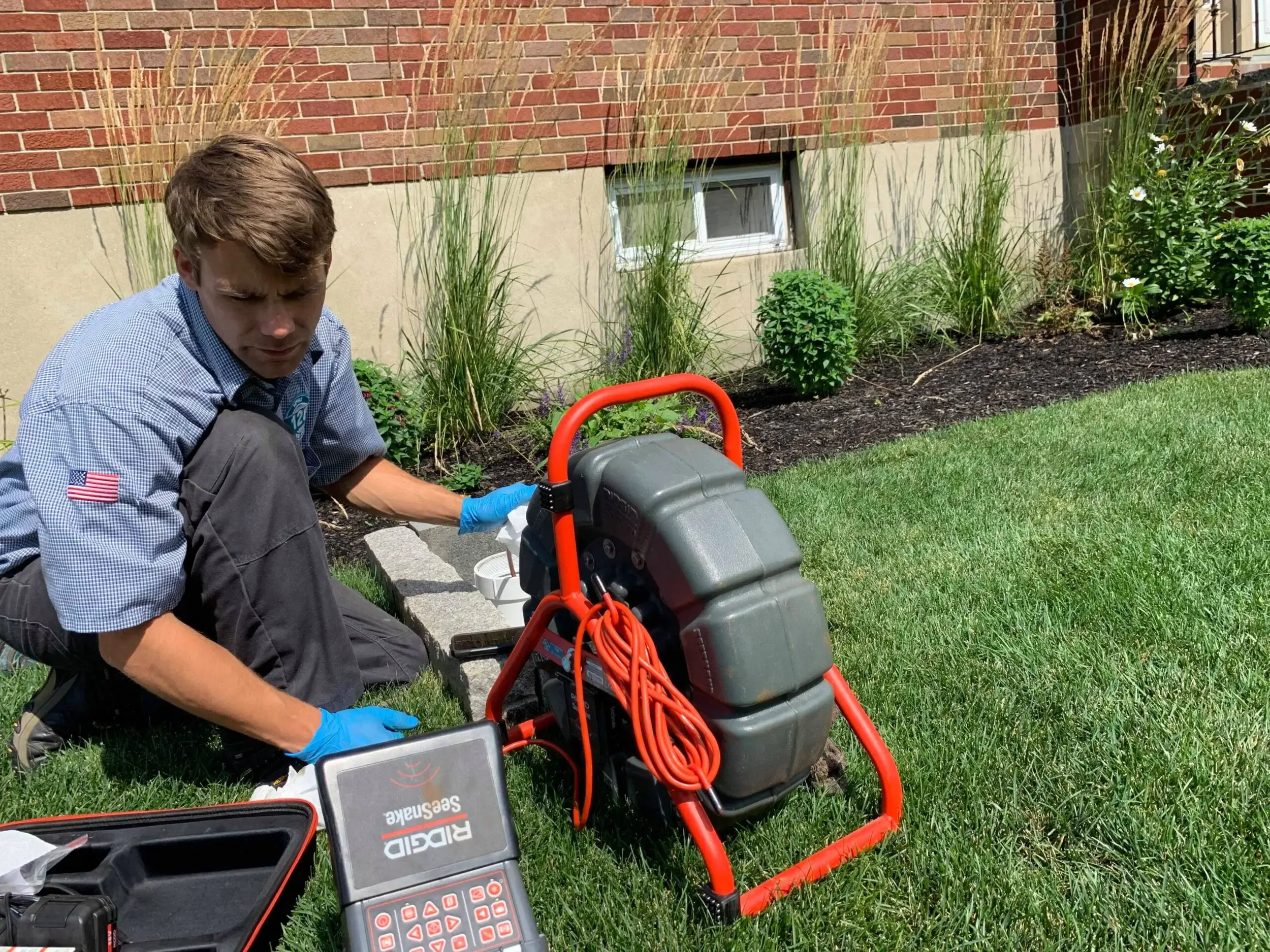 A man in a blue shirt expertly operates a Ridgid SeekTech device for drain cleaning near a brick building in Andover, carefully maneuvering across the grassy area.