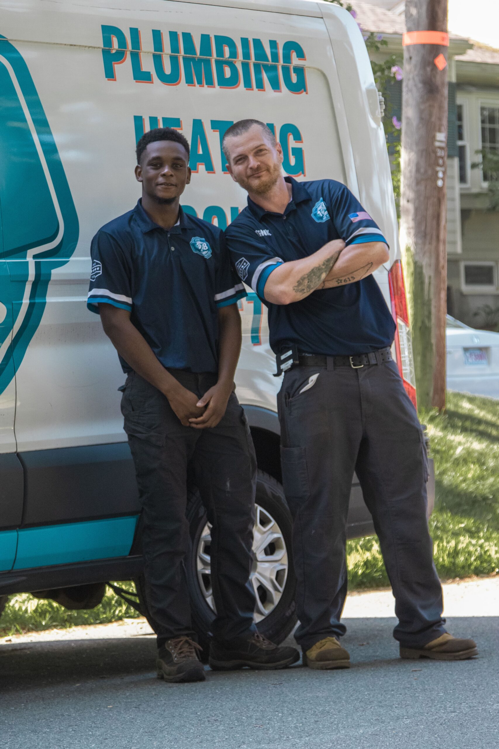 Two men in uniform stand confidently in front of a service van labeled "Plumbing Heating Cooling," ready to tackle any challenge, from a clogged drain to comprehensive repairs, right here in Boston, MA.