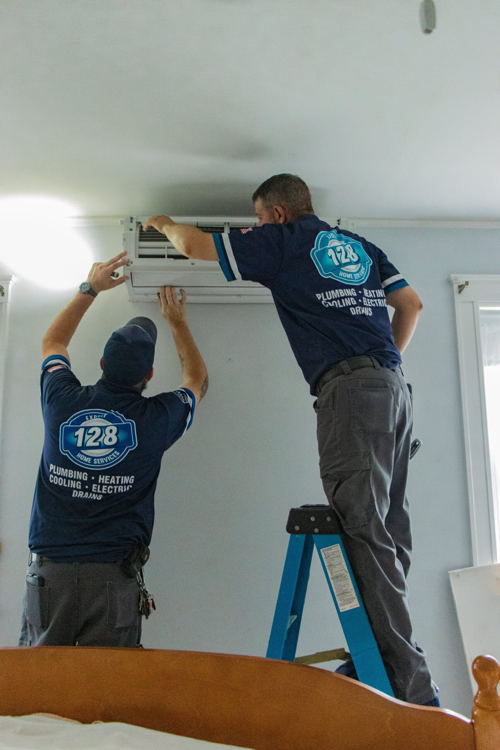 Two technicians in blue uniforms, representing Littleton Heating Services, install an air conditioning unit on a wall. One is standing on a ladder while the other assists from the ground.