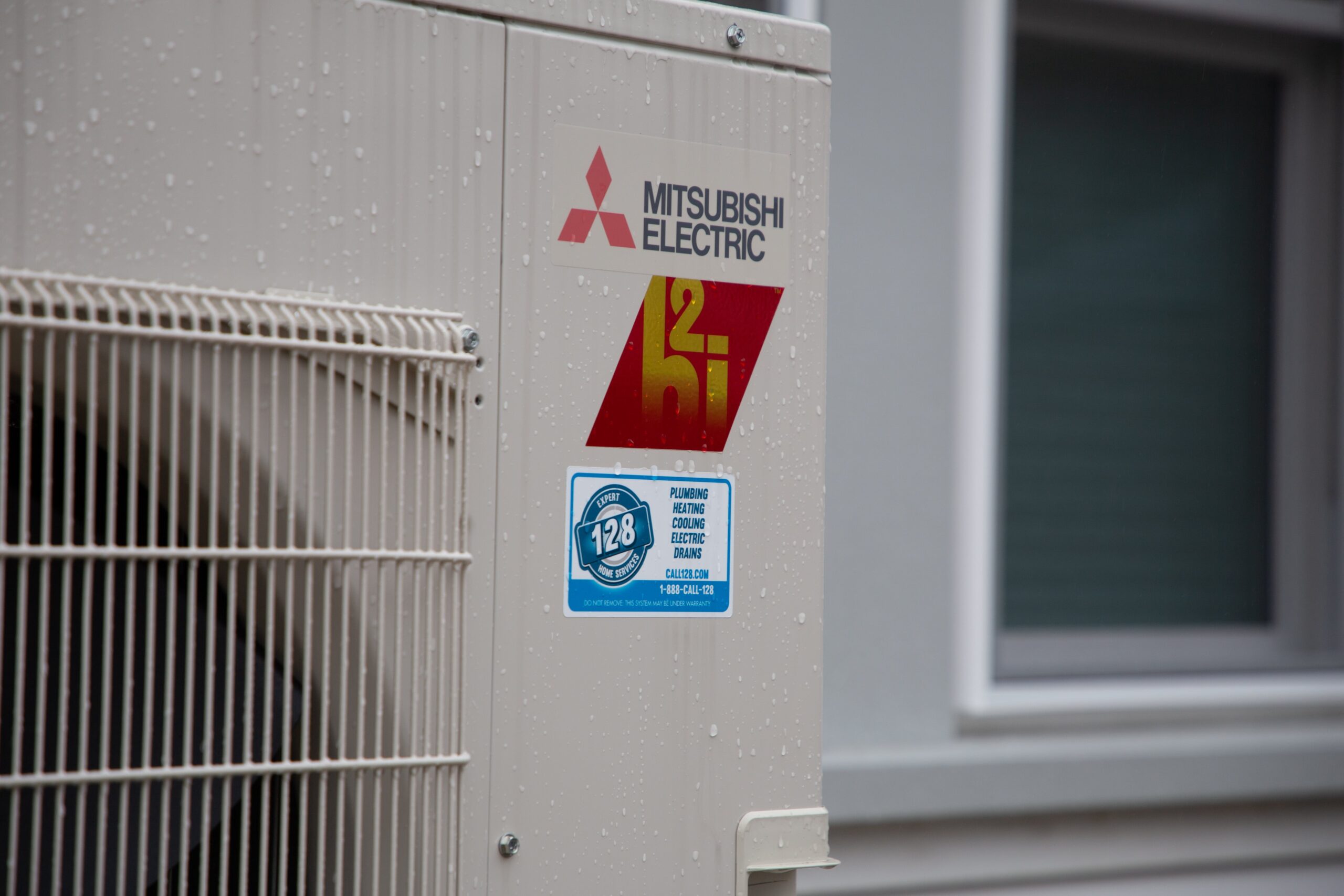 Close-up of a Mitsubishi Electric unit belonging to Westford Heating, featuring a red and yellow logo, a blue sticker, and water droplets glistening on its surface.