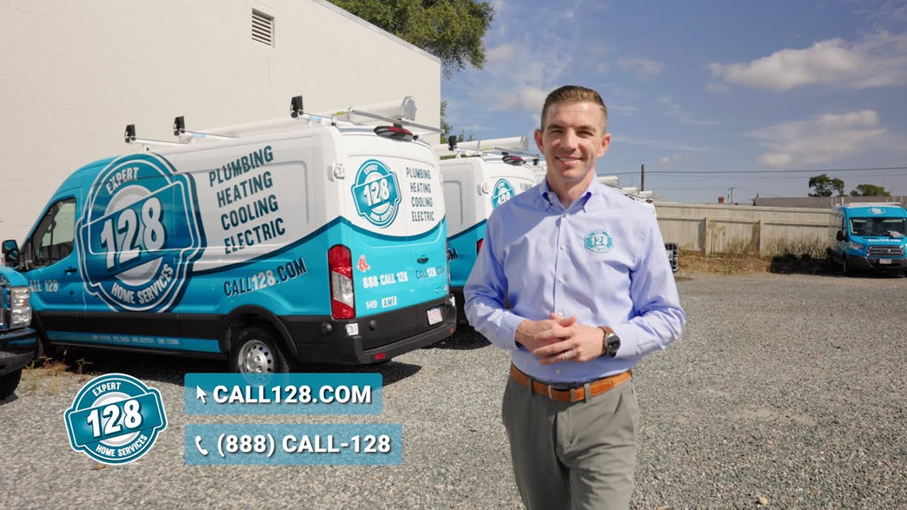 A man in uniform stands proudly next to 128 Plumbing's branded service vans in a gravel lot, showcasing logos and contact information for plumbing, heating, cooling, and electric services.
