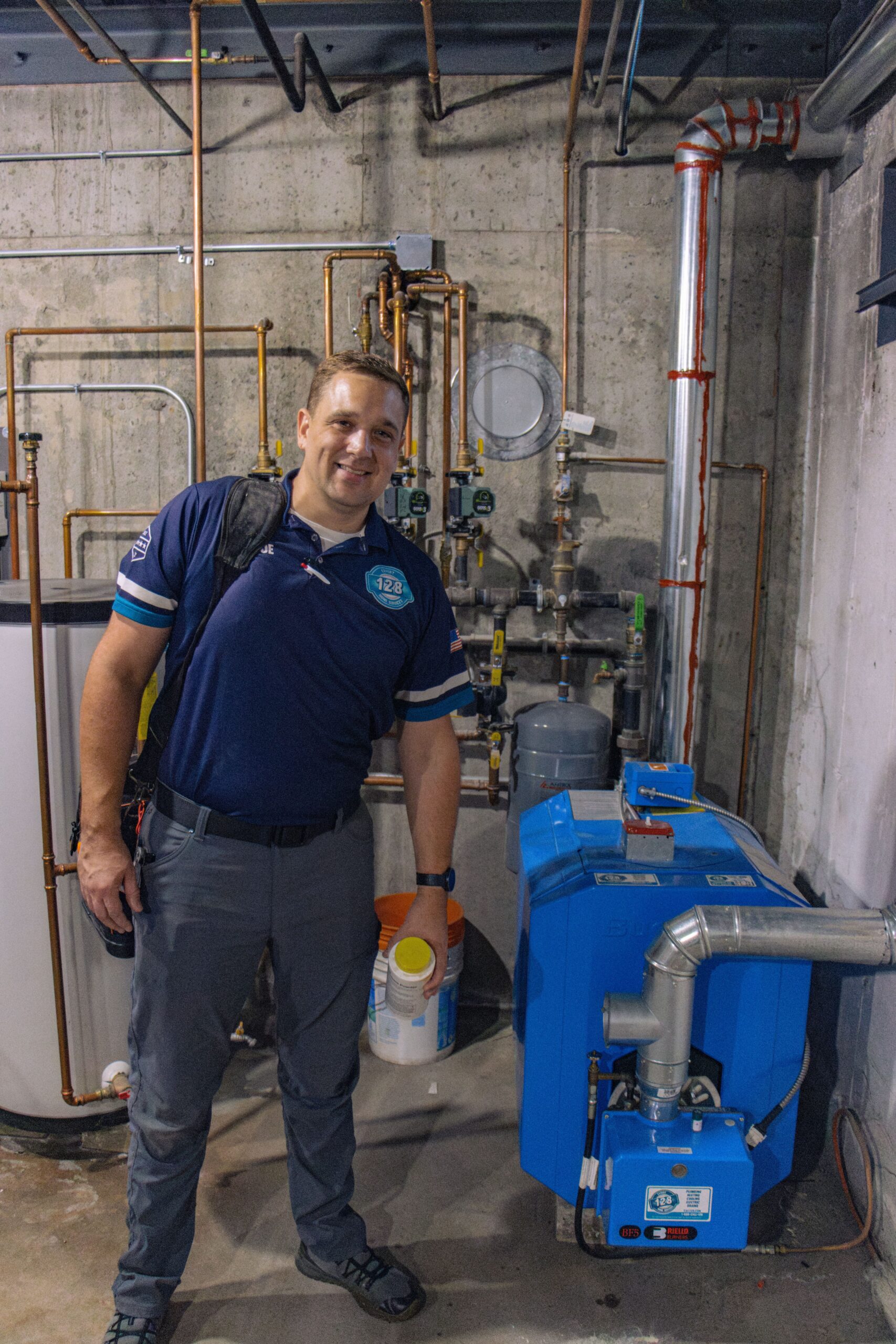 Person standing in a utility room next to blue HVAC equipment and pipes, holding a container.
