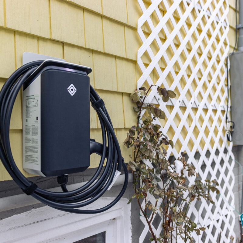 A black electric vehicle charger is mounted on the yellow exterior wall of a house, with a white lattice and some plants nearby.