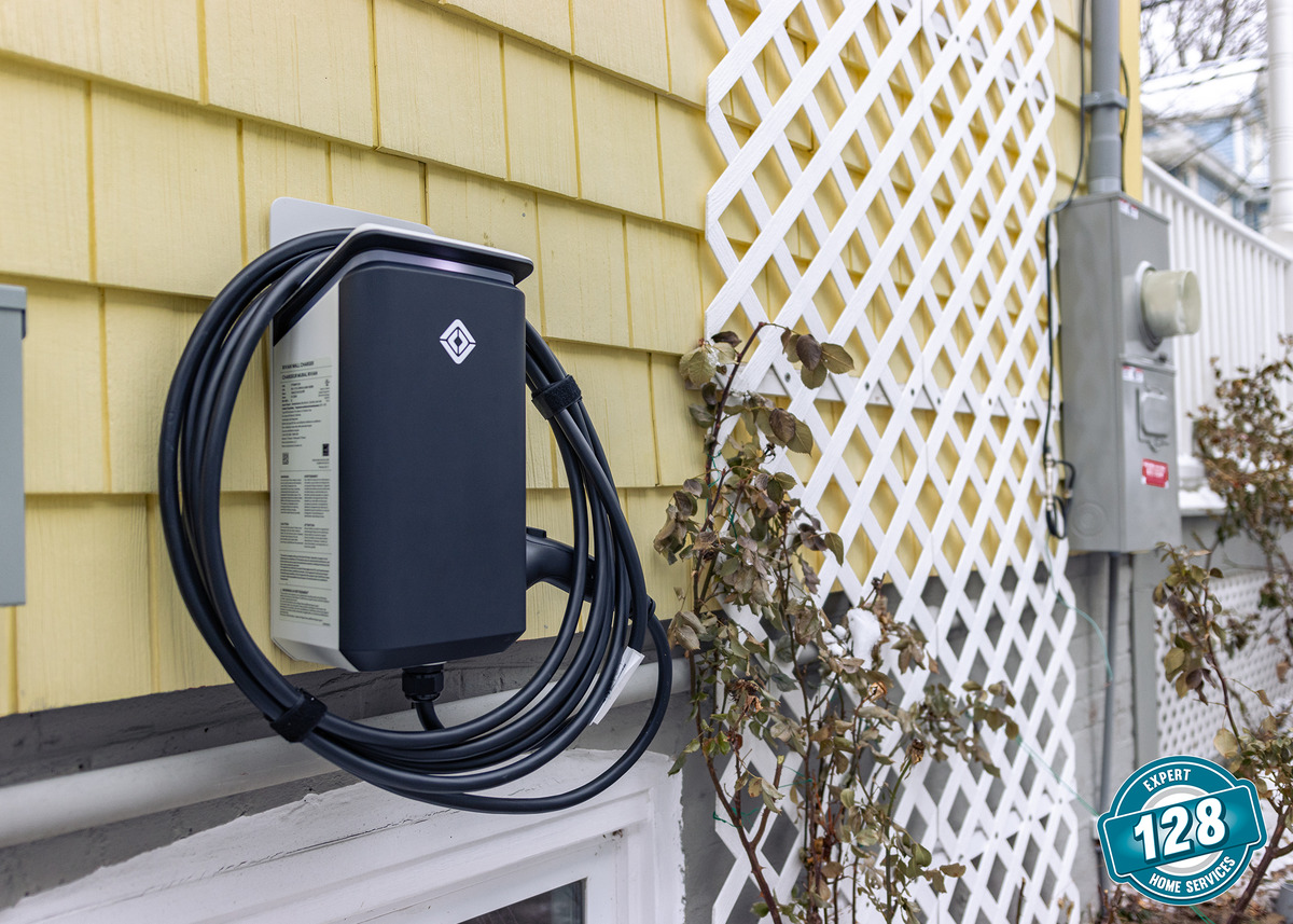 A black electric vehicle charger is mounted on the yellow exterior wall of a house, with a white lattice and some plants nearby.