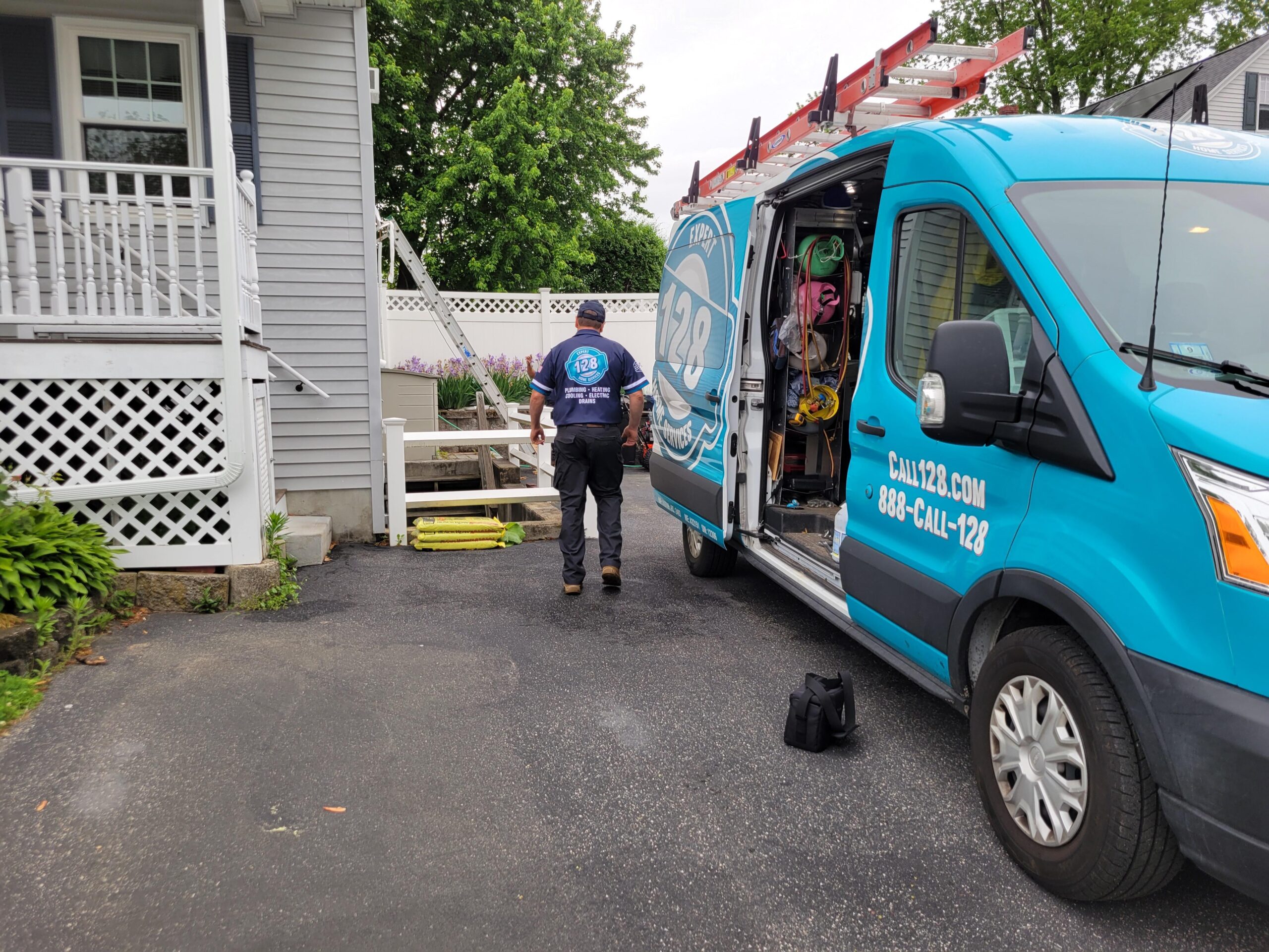 A worker from an HVAC company in Brookline MA approaches a residential staircase from a blue service van parked in the driveway. The van has a ladder on top and open side doors.