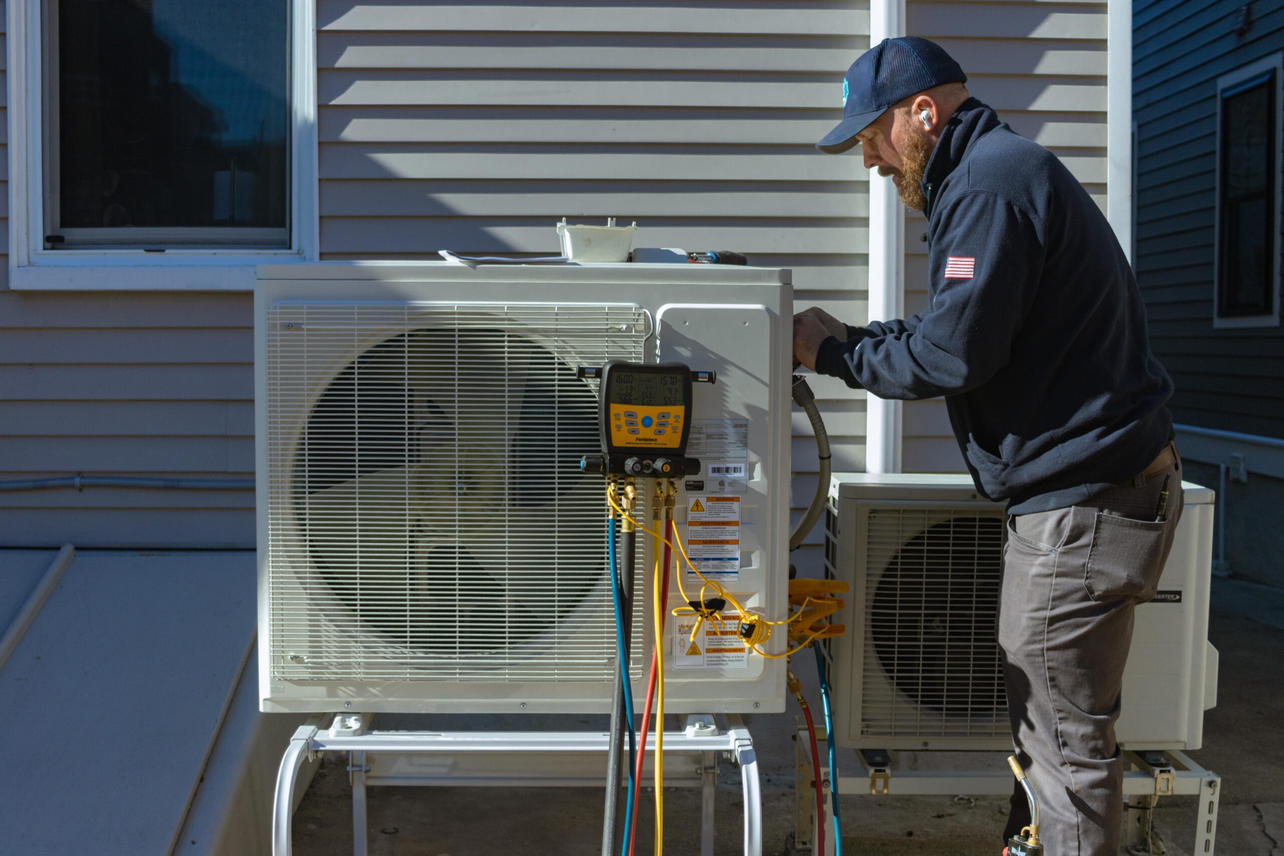 A technician from 128 Plumbing trusted HVAC company in Brighton, Boston wearing a cap and uniform, checks a residential HVAC unit outdoors, colorful hoses attached, near a house with gray siding.