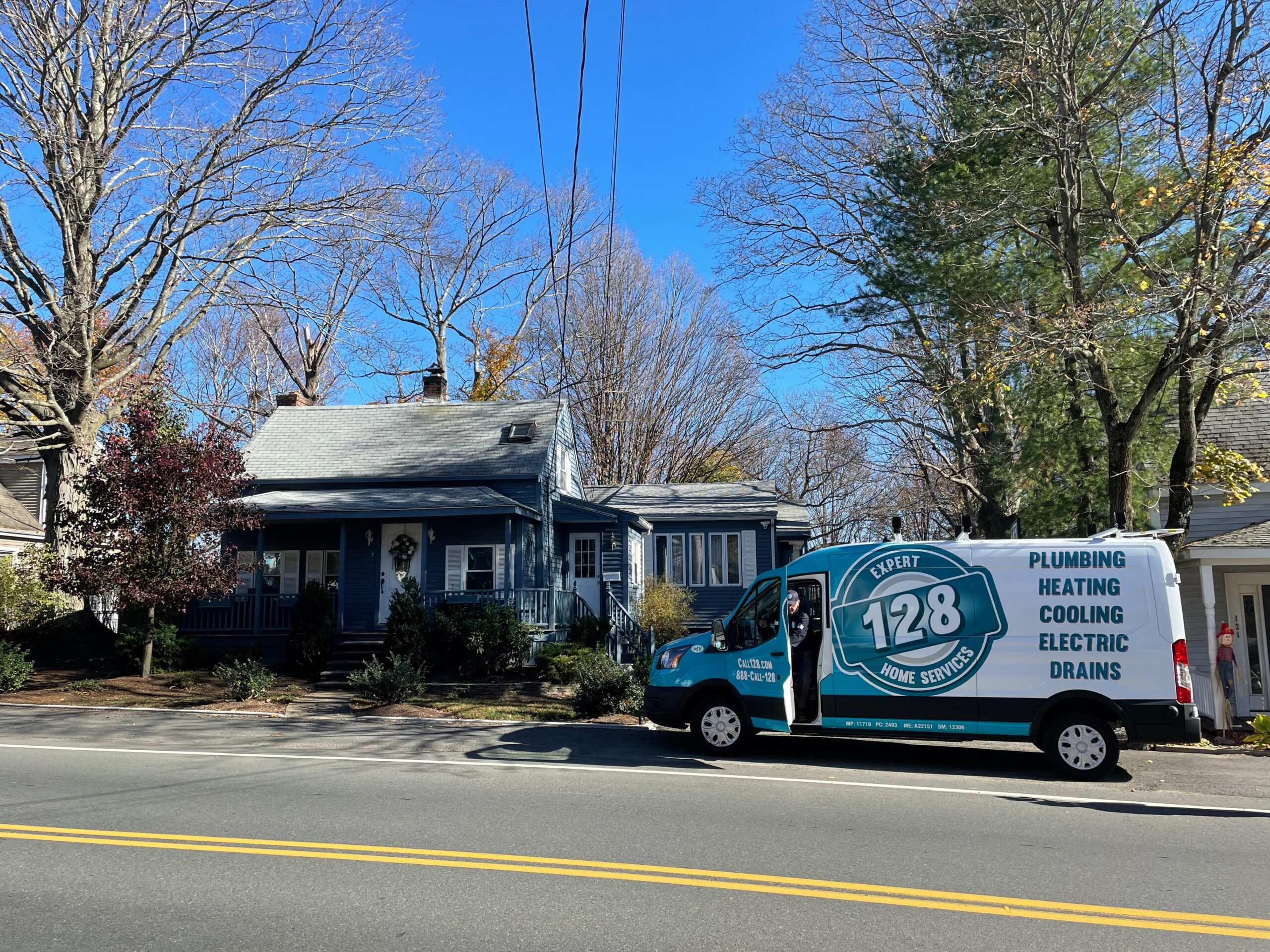 A blue service van from 128 Plumbing trusted HVAC company in back bay boston is parked on the street in front of a gray house with a porch, surrounded by bare trees on a sunny day.