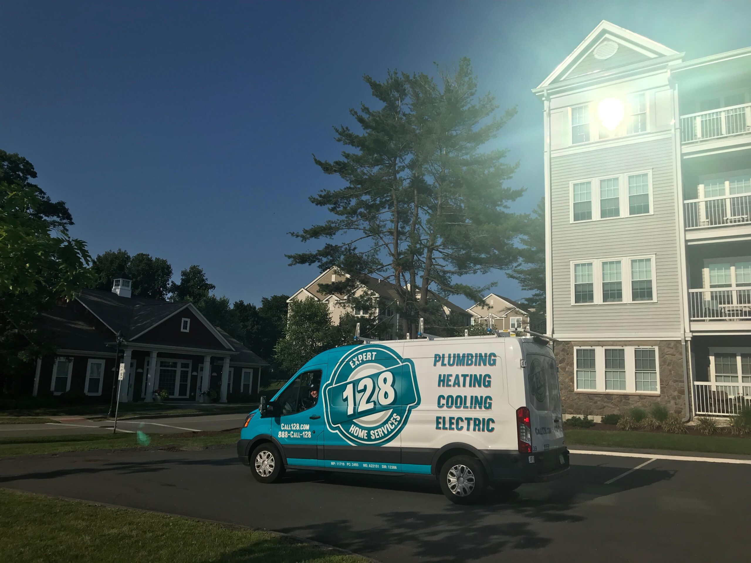 A blue service van of 128 Plubming for plumbing, heating, cooling, and electric from in Needham MA is parked near residential buildings under a clear sky performing air conditioning services.
