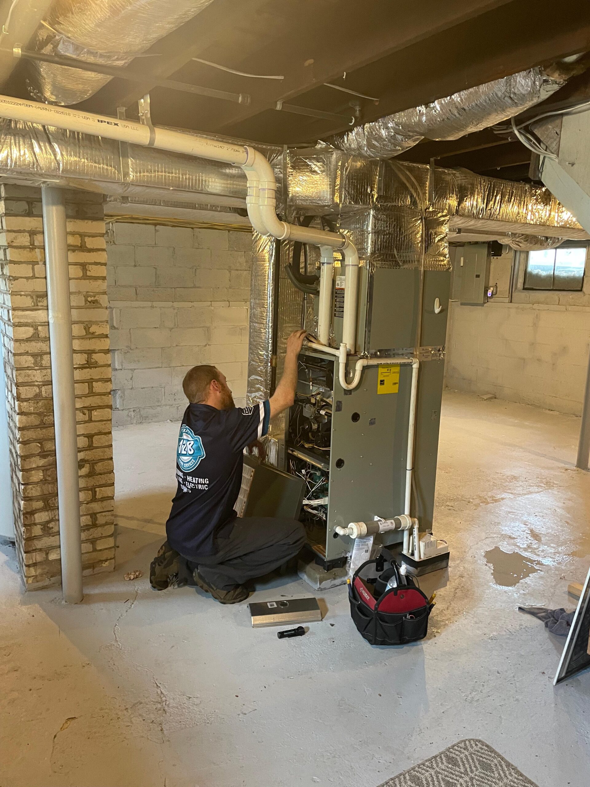 A technician in a blue uniform kneels in a Milton basement, working diligently on a furnace system surrounded by ductwork and pipes. Nearby, the logo of an air conditioning company is printed on his tool bag resting on the floor.
