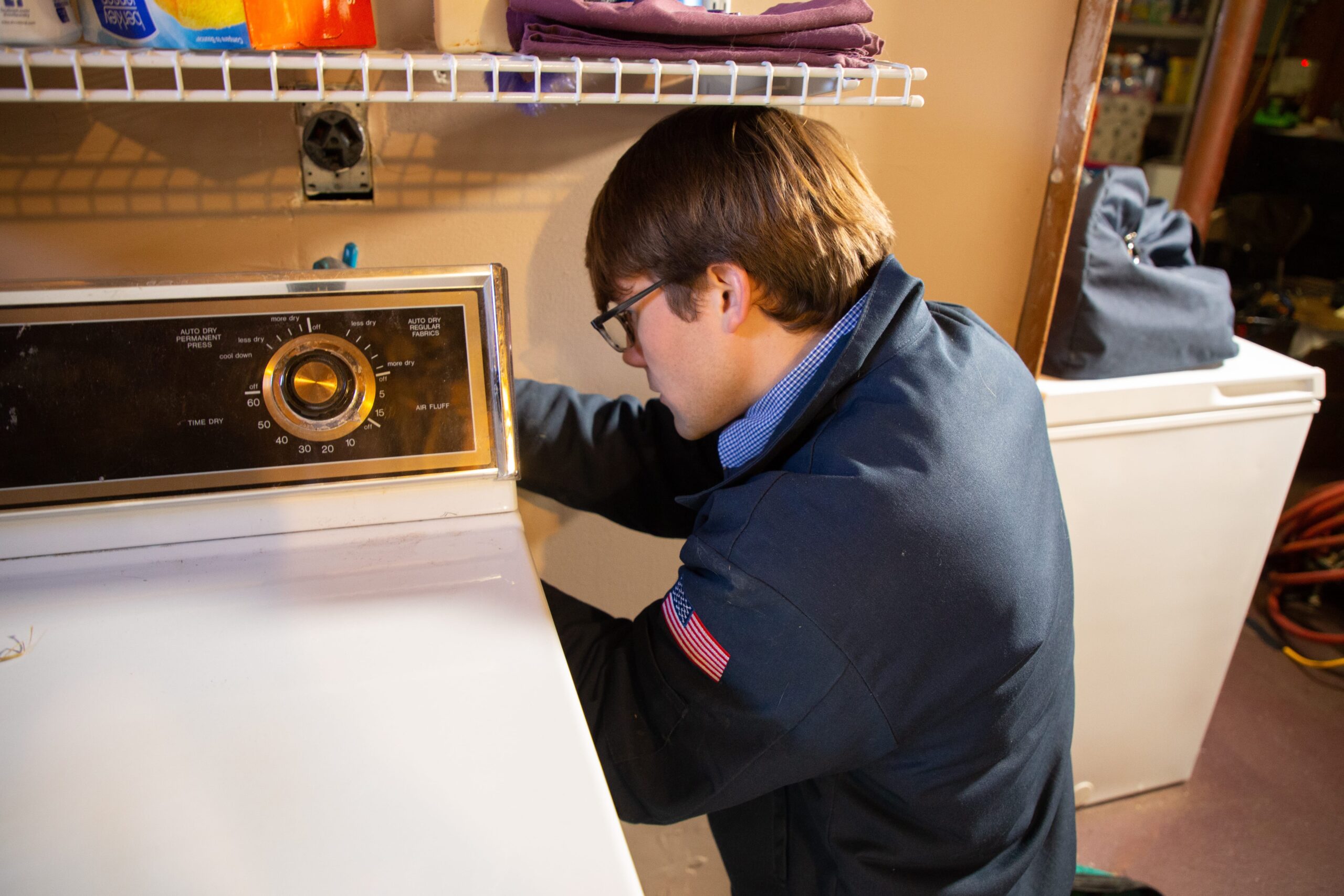 Plumber with glasses and a jacket inspecting or repairing a washing machine in a laundry room.