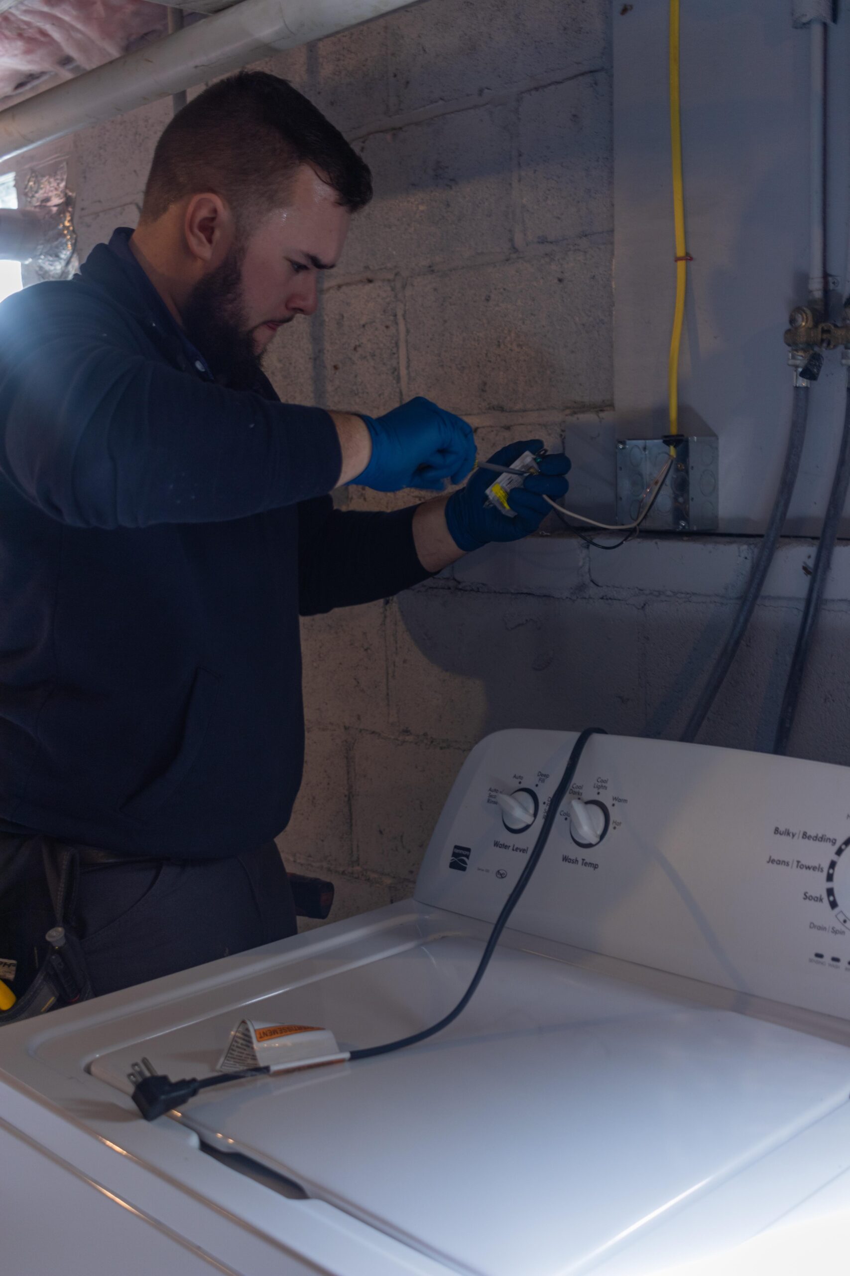 Trusted electrician from 128 Plumbing in blue gloves meticulously fixes wiring above a washing machine in a Belmont MA basement.
