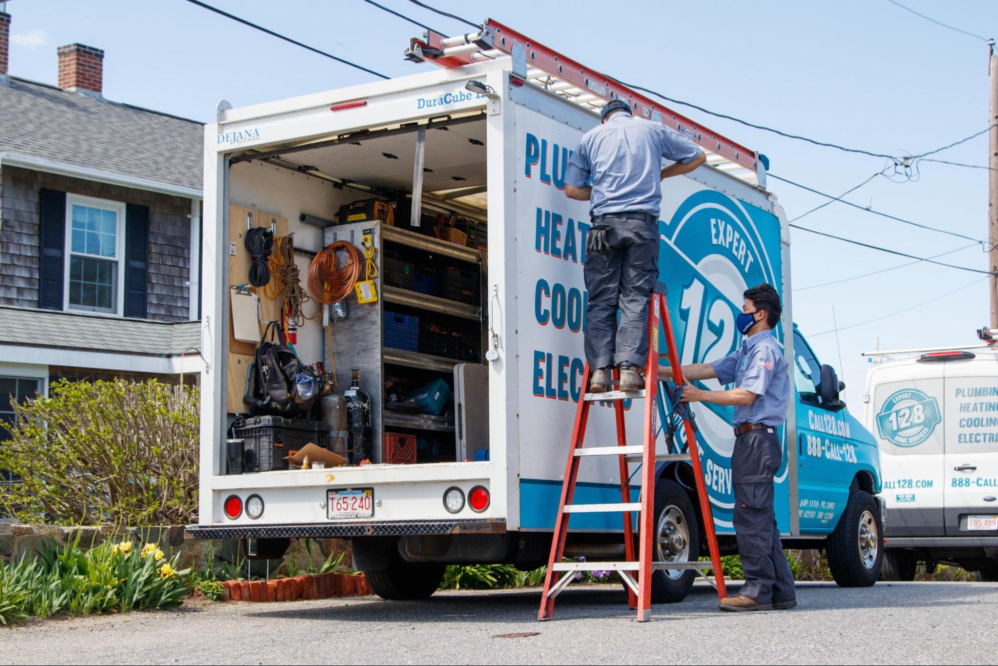 Two workers from an HVAC company in Newburyport, wearing uniforms and masks, stand by a utility truck with an open rear door, accessing equipment for plumbing and electrical services on a residential street.