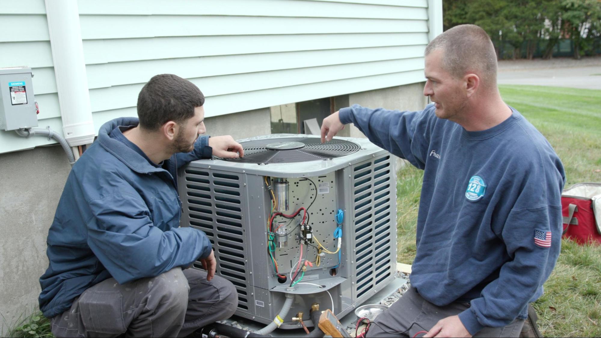 Two technicians examine and discuss the wiring of a large outdoor air conditioning unit beside a house.