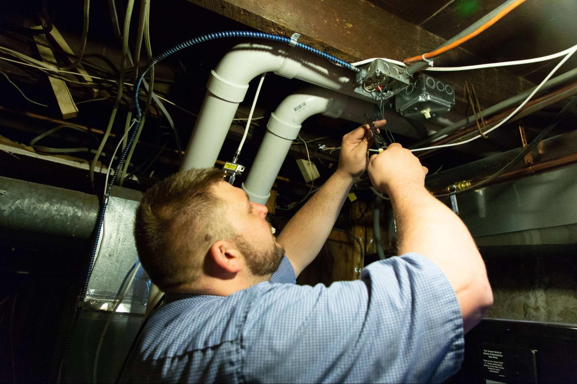 A person in a blue shirt repairs electrical wiring in a basement with exposed pipes and cables.