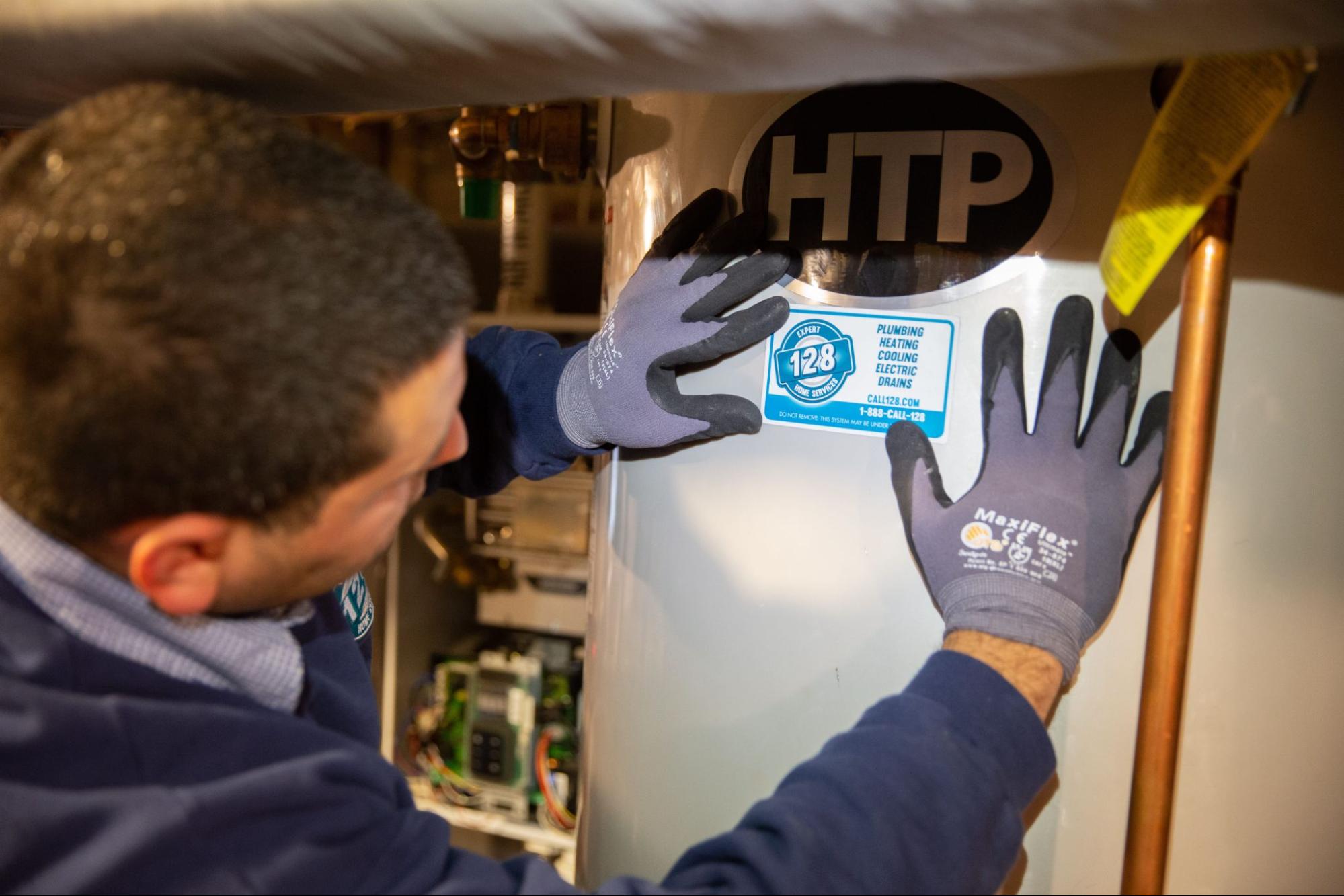A person wearing gloves is placing a sticker on a water heater tank. The sticker displays information about plumbing, heating, and cooling services with contact details.