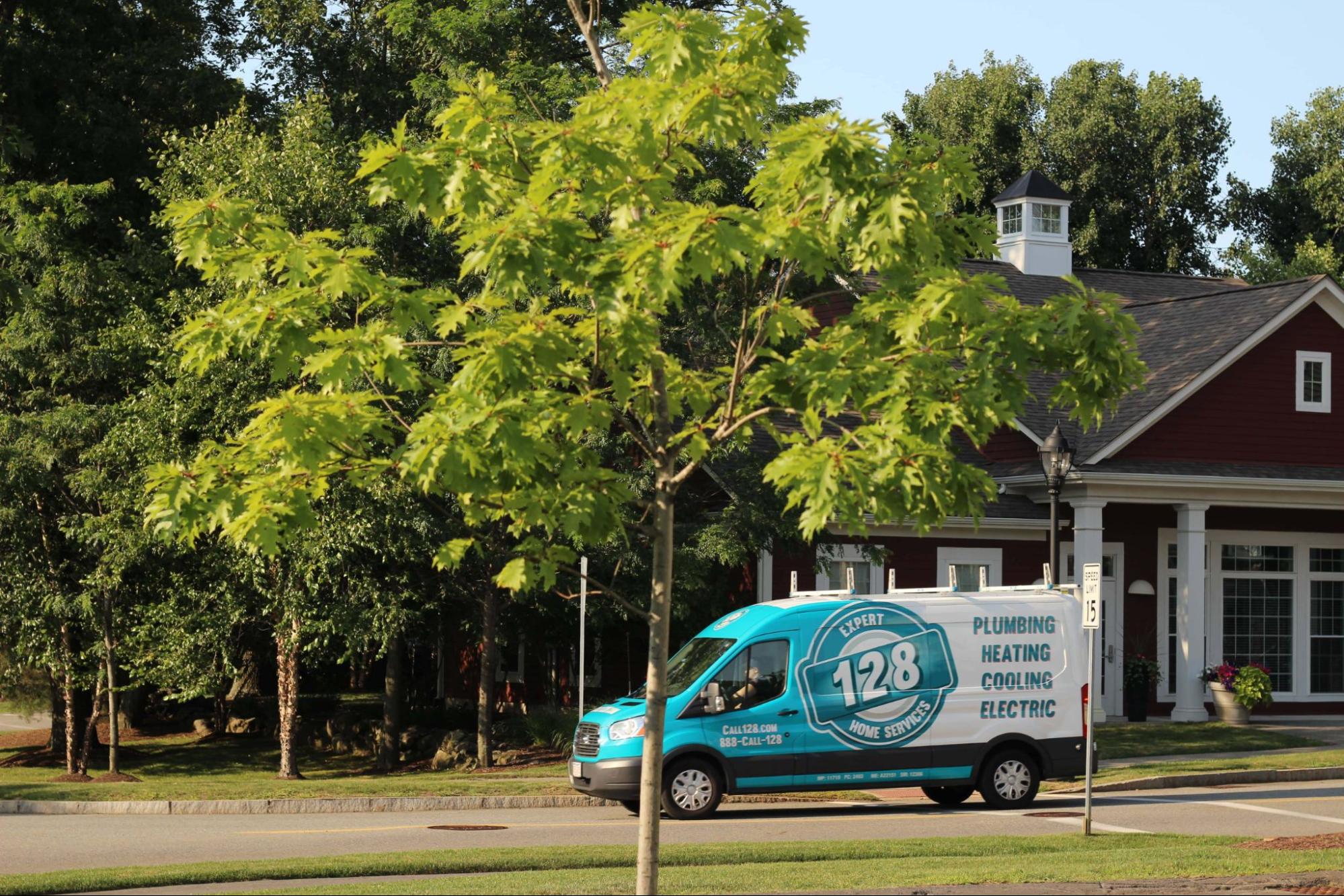 A teal van with company branding and text for plumbing, heating, cooling, and electric services is parked on a suburban street near a tree and a house.