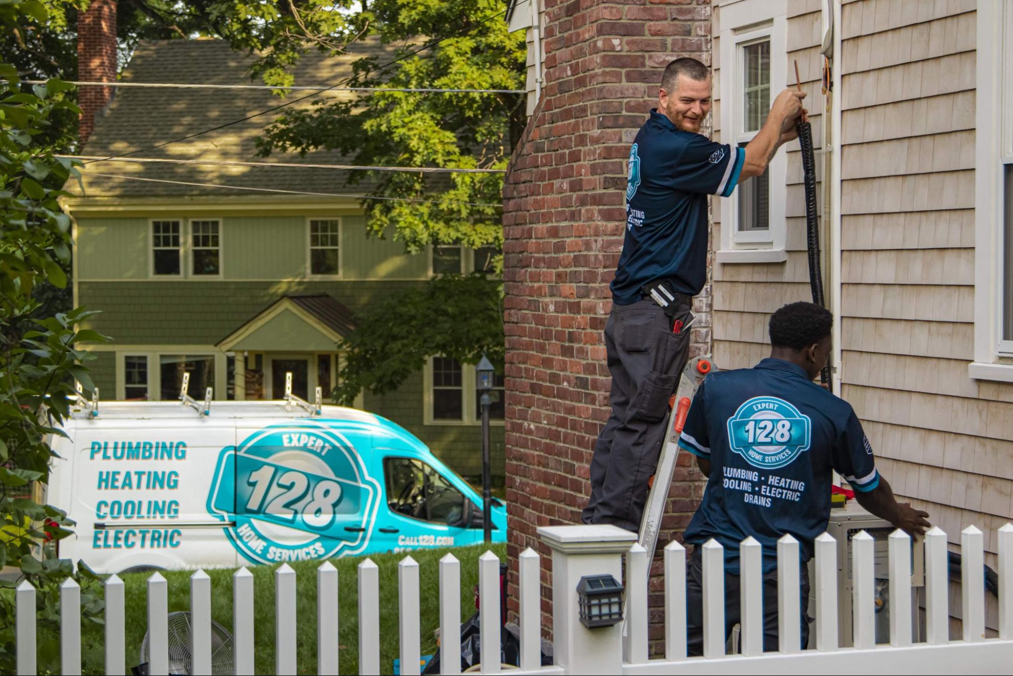 Two workers from "128 Expert Services" wearing blue uniforms perform maintenance on the side of a house. A company van with "Plumbing, Heating, Cooling, Electric" is parked nearby.
