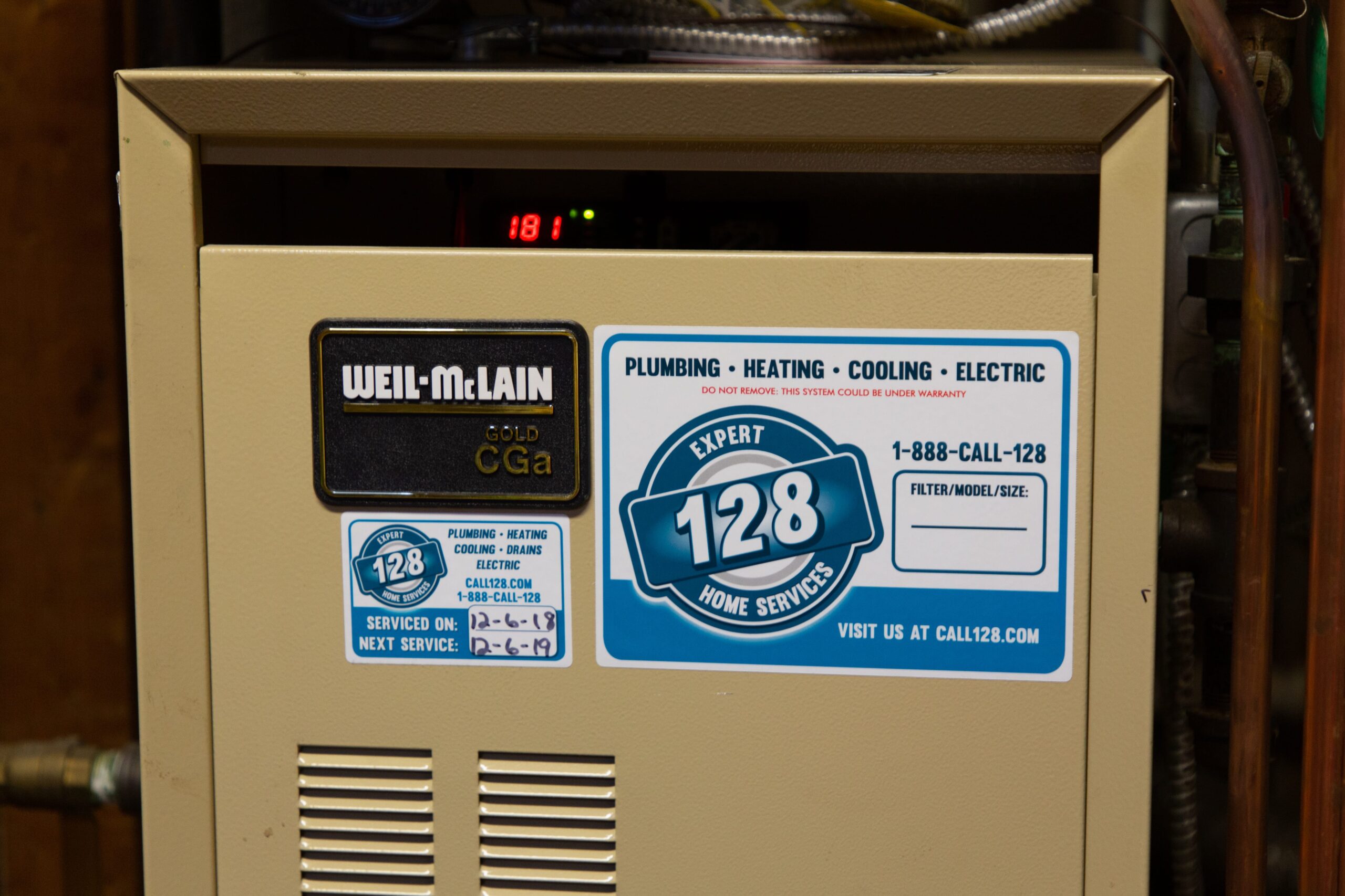 Front of a Weil-McLain Gold CGa boiler with a sticker displaying contact information for a plumbing and heating service company, including phone number and website.