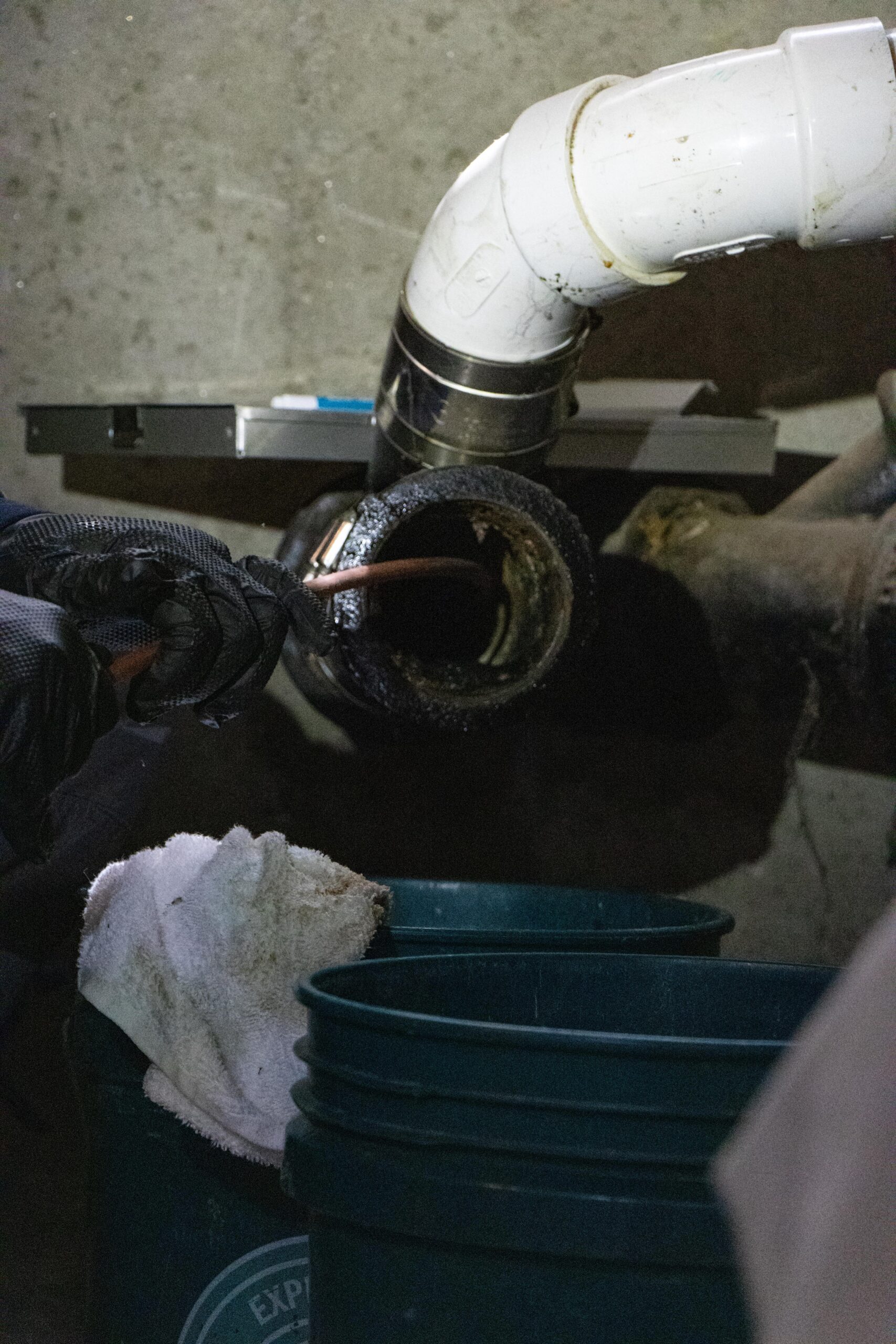 A Somerville plumber is using a wrench to work on a grimy pipe, surrounded by a white cloth and green buckets on the concrete floor.