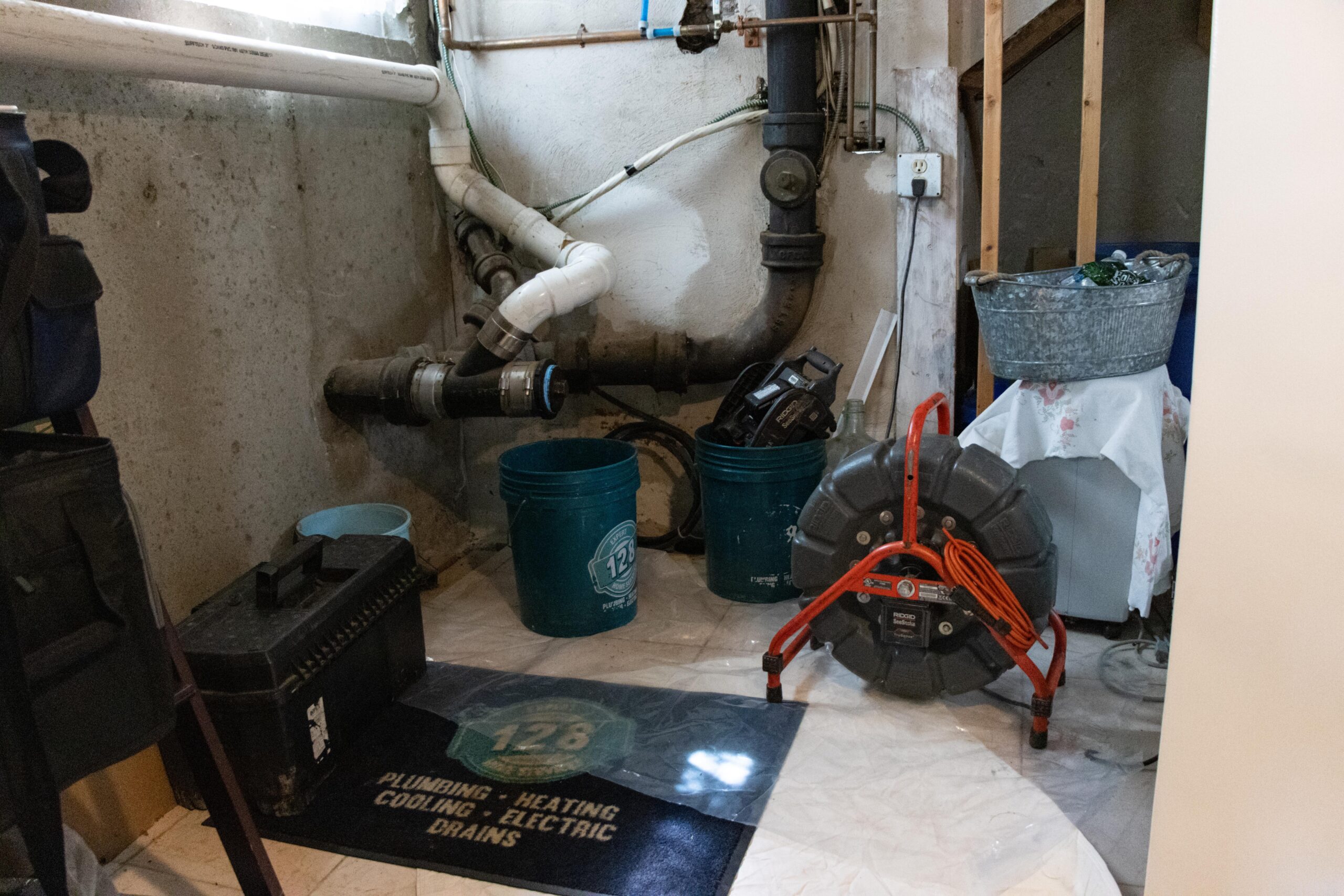 In the basement corner, plumbing pipes and a toolbox accompany a large drain cleaning machine on a mat labeled "plumbing, heating, cooling, electric, drains." This setup is typical for an HVAC company for plumber in Brighton MA.