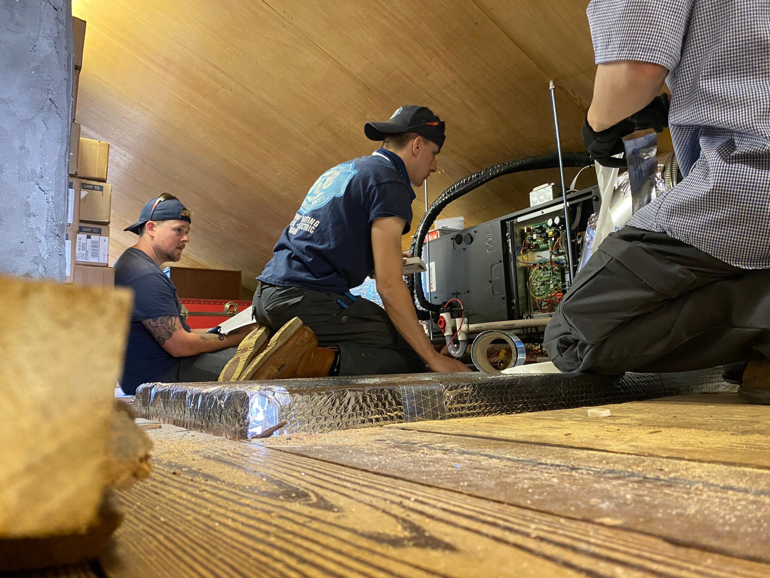 Three team members from an air conditioning company in South End Boston expertly install HVAC equipment in an attic, with wooden flooring and exposed beams adding rustic charm to their workspace.