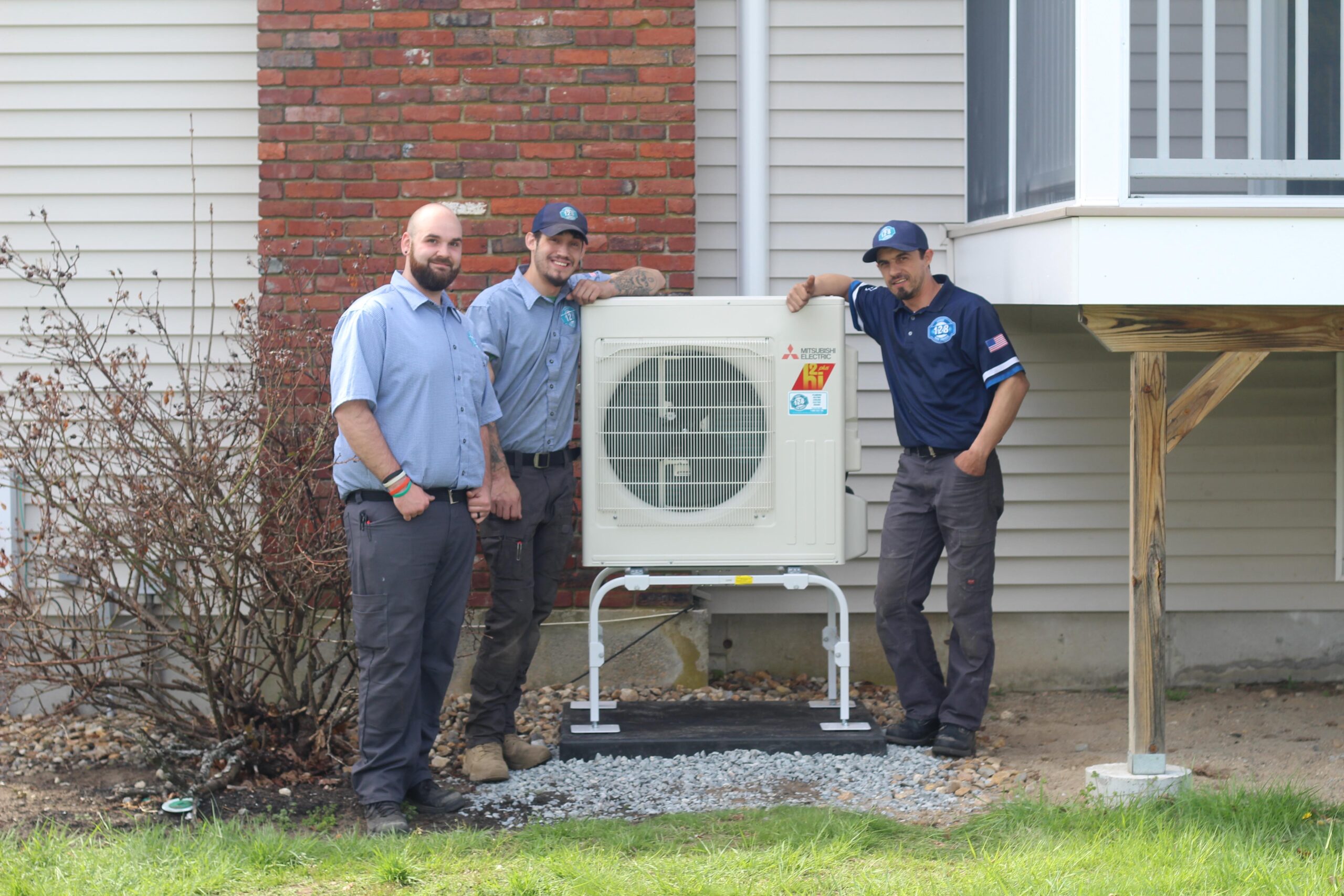 Three uniformed men from an HVAC company in Beacon Hill MA stand beside an installed outdoor unit against a house, featuring a brick and siding exterior.