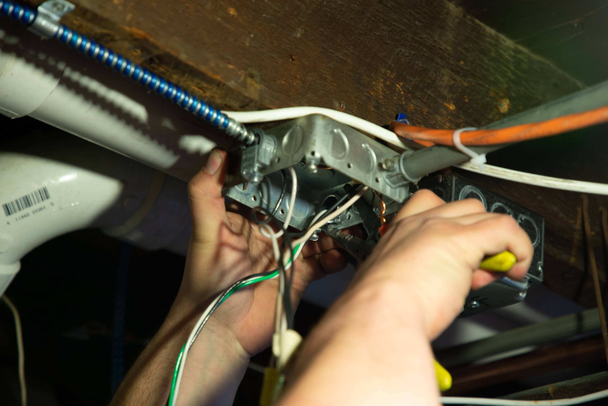 An electrician in Brighton MA precisely adjusts wiring, skillfully using tools on a metal junction box beneath a wooden beam.
