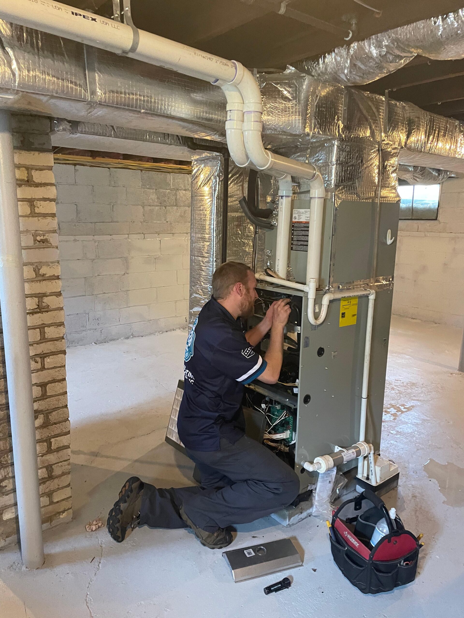 A technician from an HVAC company in Somerville kneels amidst a maze of ducts and tools, expertly fixing a furnace in the basement.
