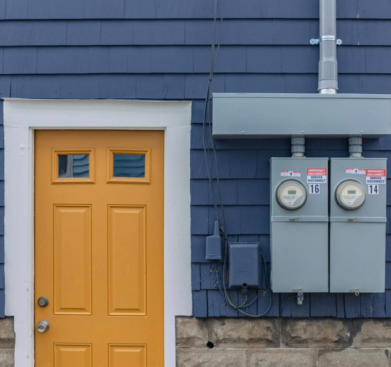 A yellow door next to two electric meters mounted on a blue exterior wall with visible wiring and utility boxes.
