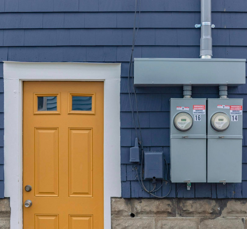 A yellow door next to two electric meters mounted on a blue exterior wall with visible wiring and utility boxes.