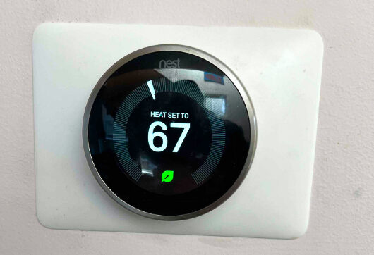 A Nest thermostat mounted on a white wall displays the heat set to 67 degrees Fahrenheit with an energy-saving leaf icon.