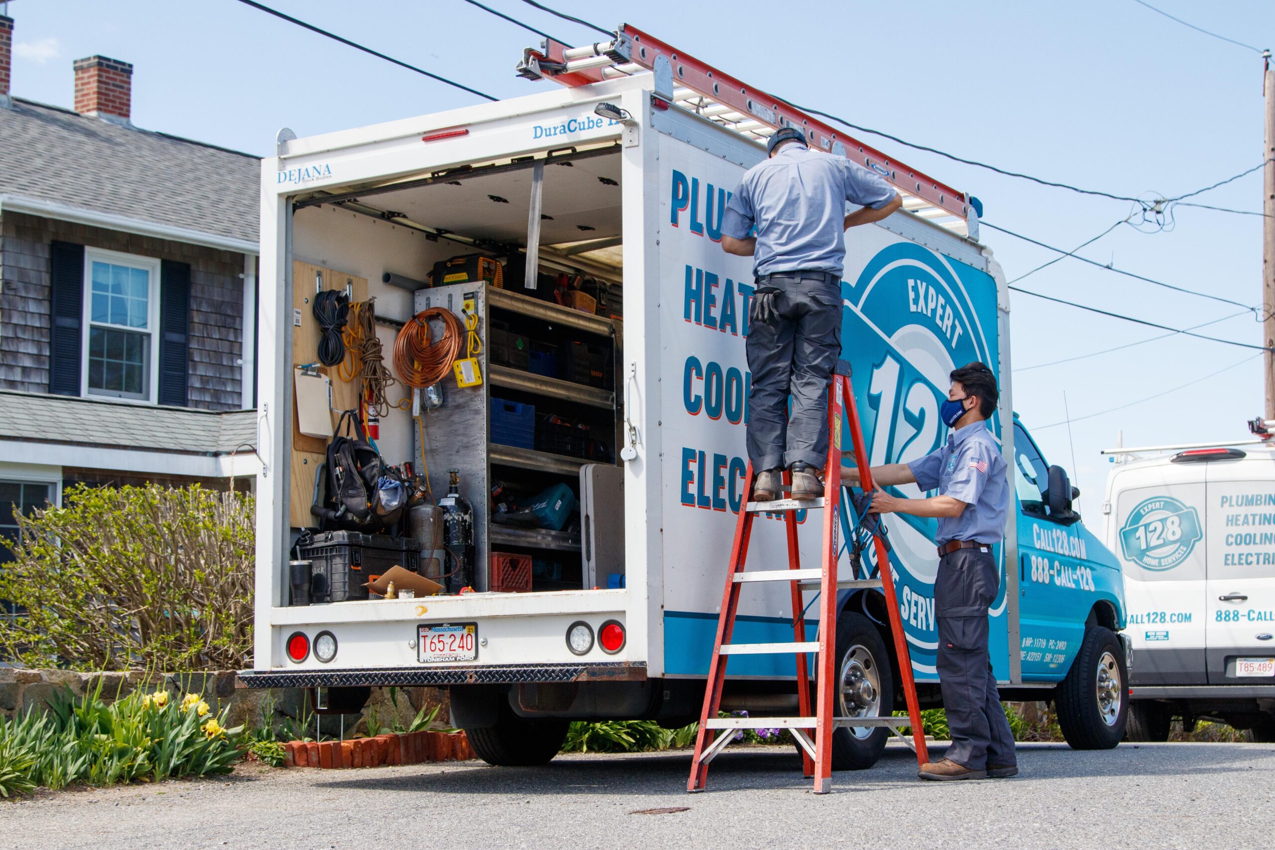 Two workers stand by an open utility van with tools and supplies, one on a ladder working near the roof, the other beside the van, handling an air conditioning replacement in Boston.