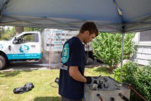 A technician in a blue shirt works with gauges and tools under a tent outside, providing drain cleaning service Milton, with a company truck and green bushes in the background.