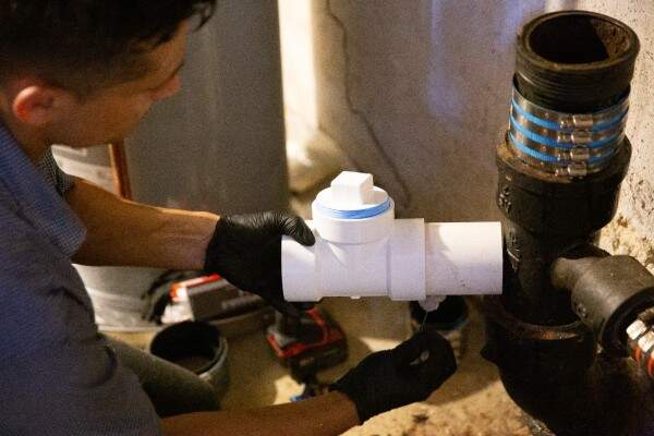 A person wearing black gloves installs a white PVC check valve onto a black metal pipe in a basement, highlighting skilled drain cleaning services Needham residents can rely on.