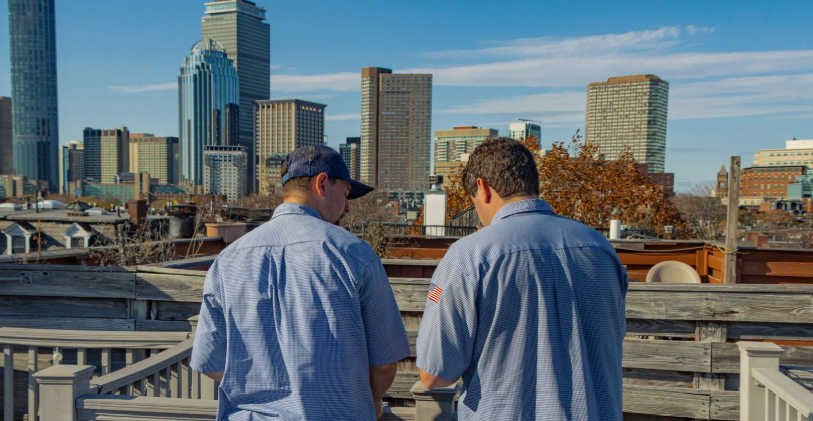 Two men in uniform shirts stand on a rooftop deck, facing away from the camera, with a city skyline visible in the background under a clear sky.