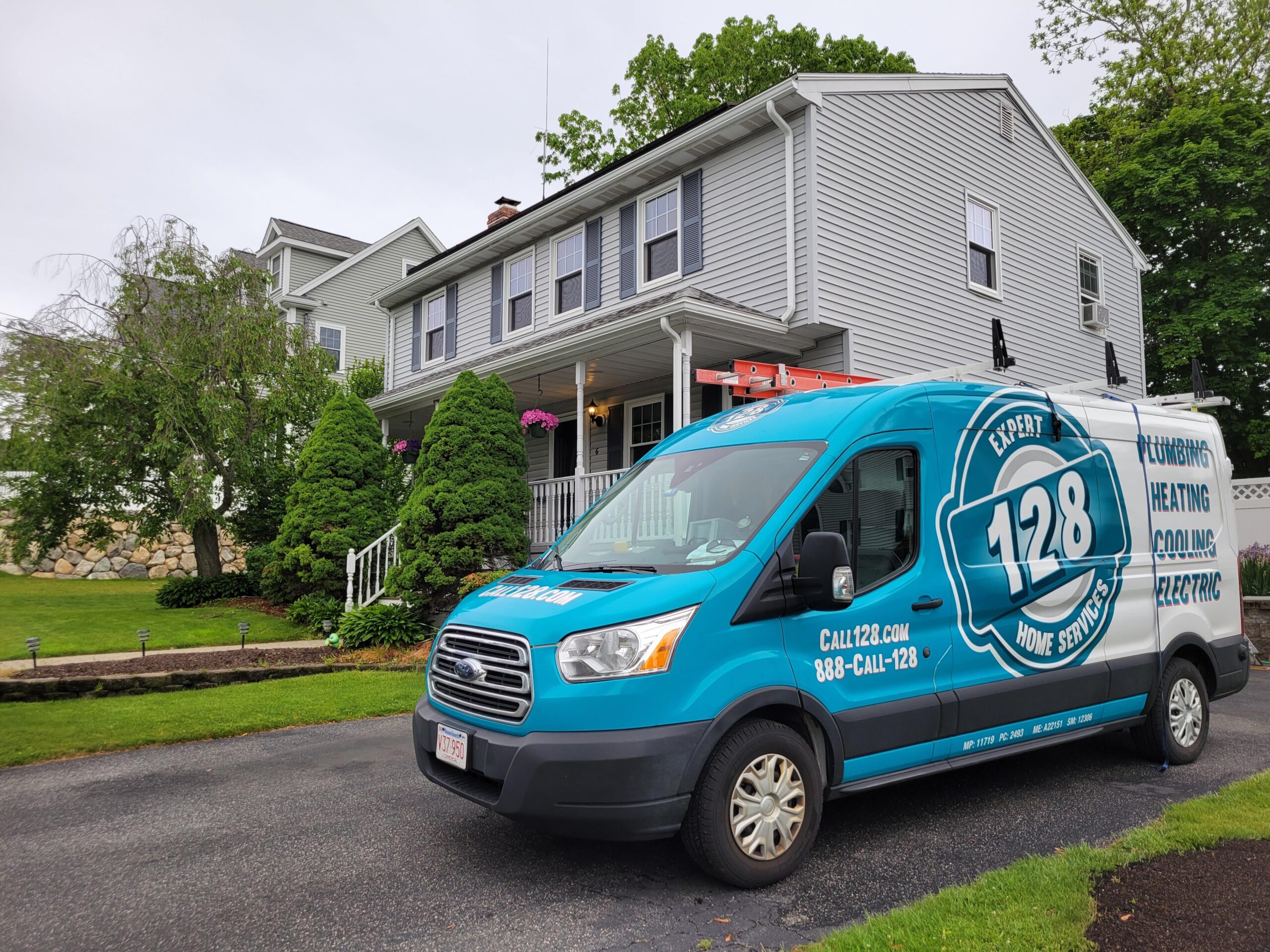 A blue 128 Home Service van, belonging to a trusted HVAC company in Medford, is parked in the driveway of a two-story gray house with white trim and a well-kept yard.